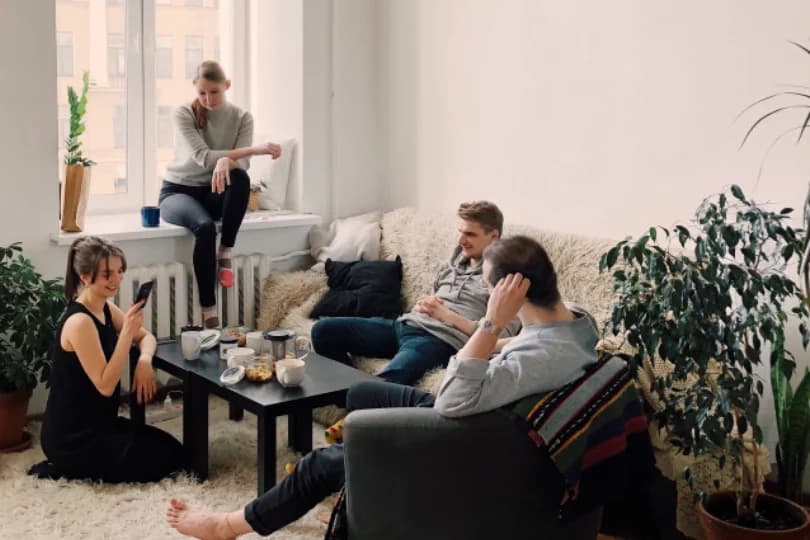 Four friends relaxing in a bright living room, one sitting on the windowsill and one checking her phone on the rug