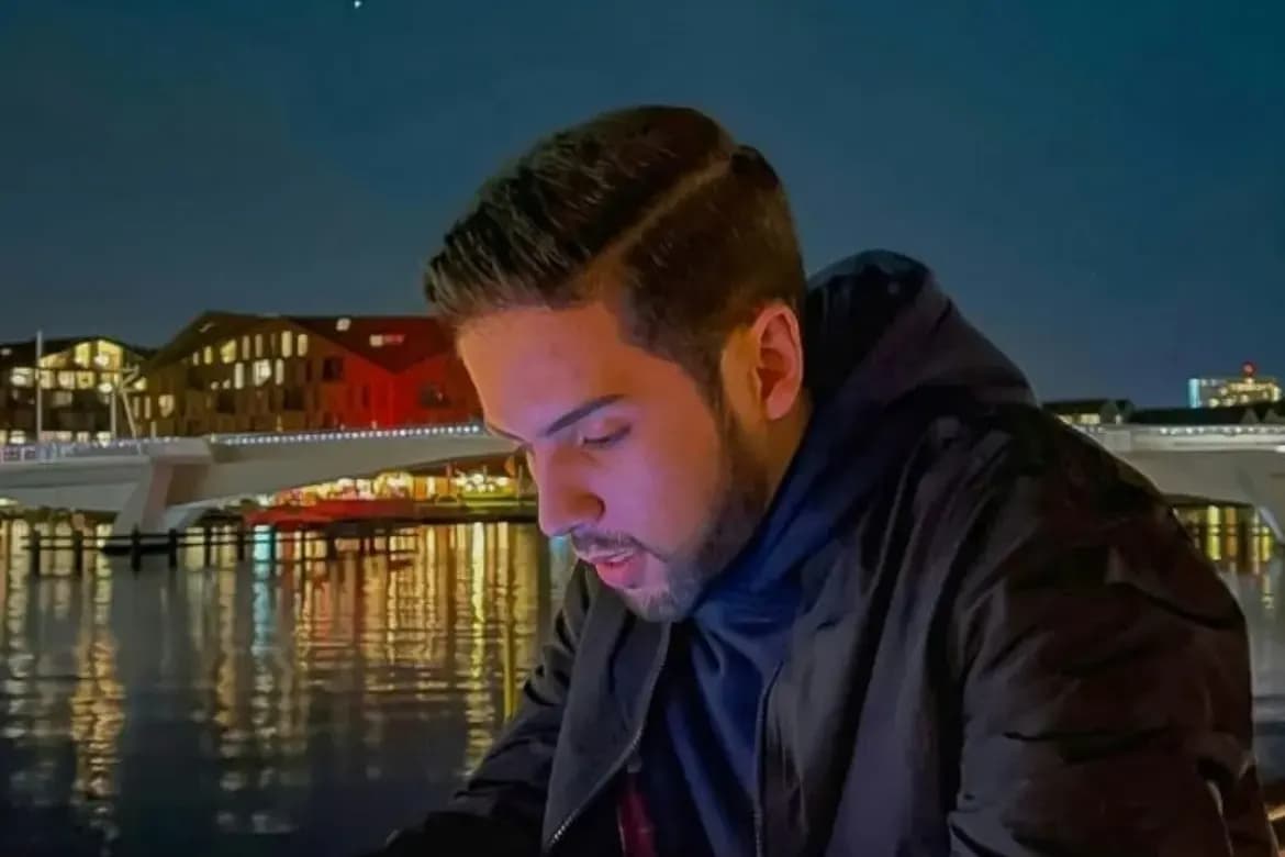 Ein Mann sitzt abends am Wasser und schaut auf sein Smartphone, im Hintergrund eine beleuchtete Brücke und Stadtlichter