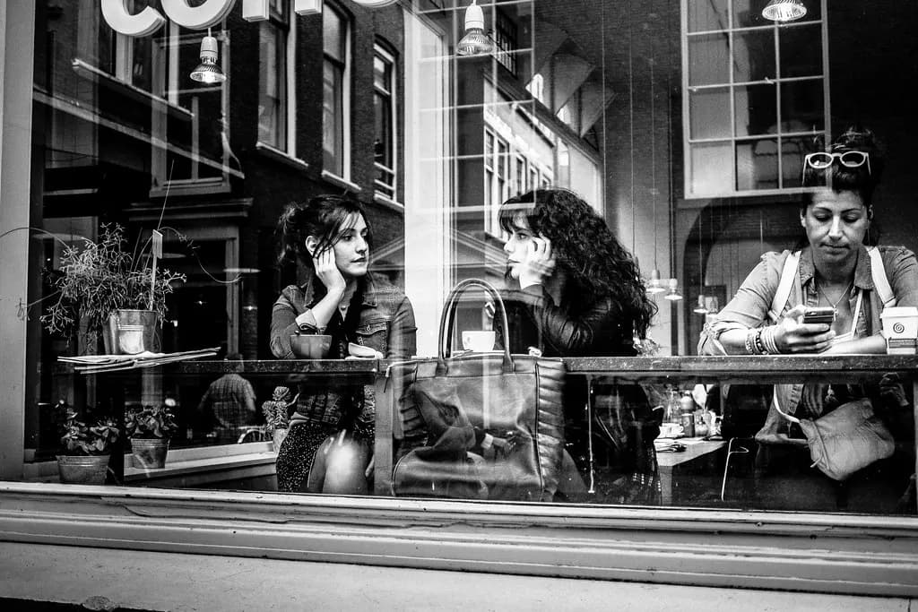 Zwei Frauen sitzen am Fenster eines Cafés und unterhalten sich, fotografiert durch die Fensterscheibe in Schwarz-Weiß