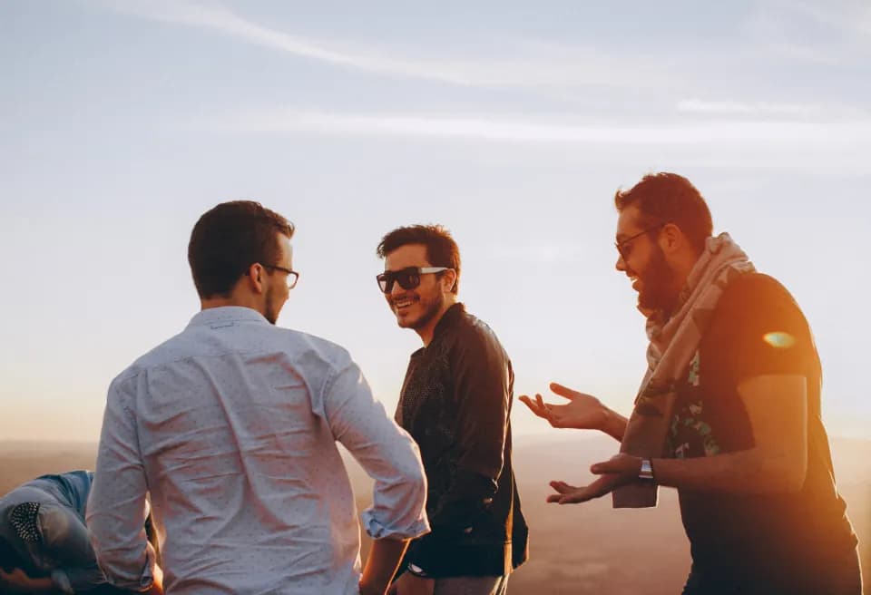 Drei Freunde stehen im warmen Abendlicht auf einer Anhöhe, reden und lachen miteinander.