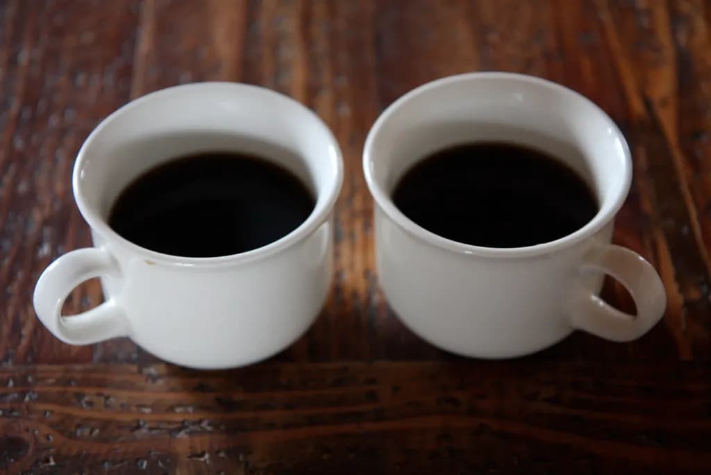 Zwei weiße Tassen mit schwarzem Kaffee stehen nebeneinander auf einem dunklen Holztisch